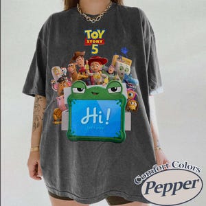 May include: A charcoal gray t-shirt featuring a graphic print of Toy Story 5 characters surrounding a green frog-shaped tablet with the text Hi! Let's play! The shirt has a vintage-washed appearance and a crew neckline with short sleeves.