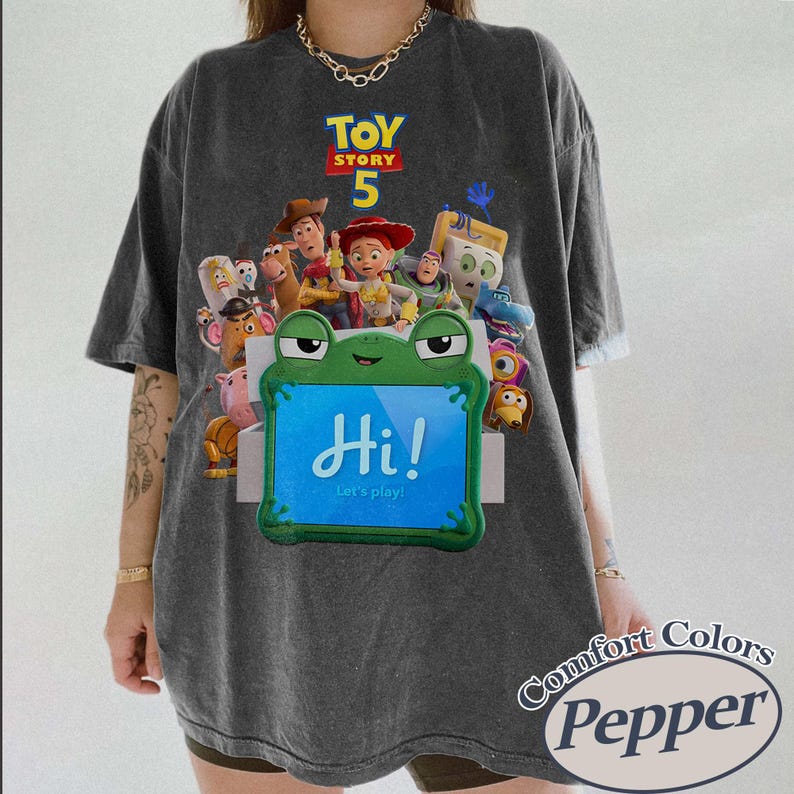 May include: A charcoal gray t-shirt featuring a graphic print of Toy Story 5 characters surrounding a green frog-shaped tablet with the text Hi! Let's play! The shirt has a vintage-washed appearance and a crew neckline with short sleeves.