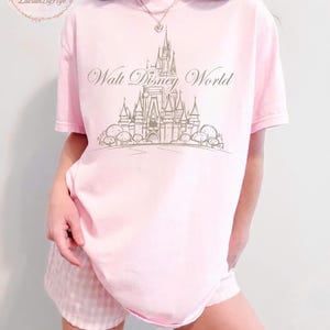 May include: A light pink t-shirt with the words "Walt Disney World" and a castle graphic in a light brown color. The shirt is paired with pink and white checkered shorts. The shirt has short sleeves.