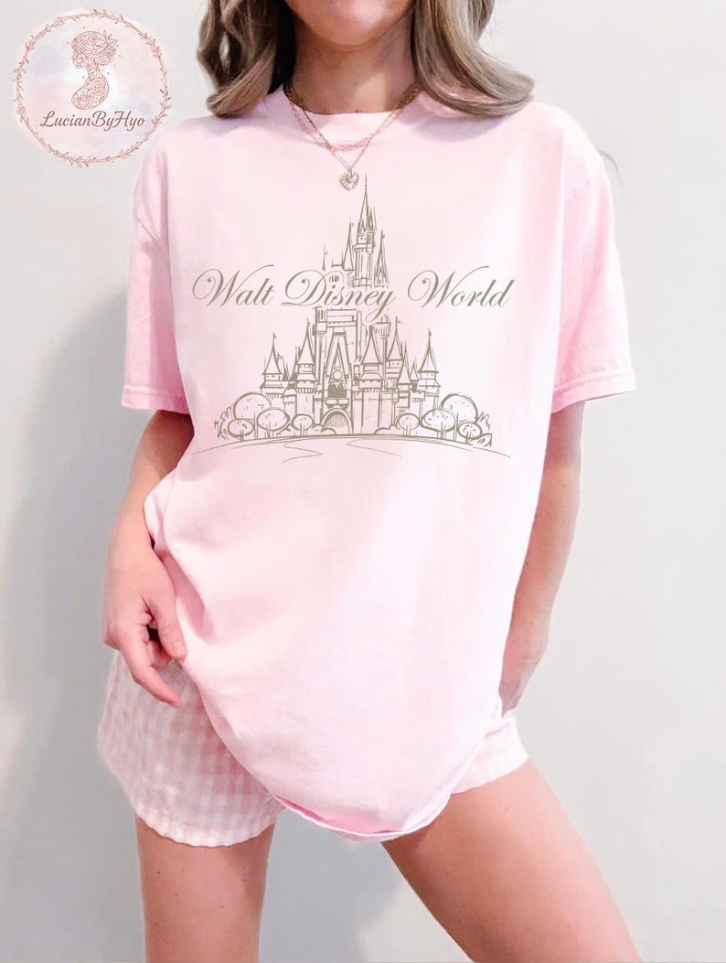 May include: A light pink t-shirt with the words "Walt Disney World" and a castle graphic in a light brown color. The shirt is paired with pink and white checkered shorts. The shirt has short sleeves.