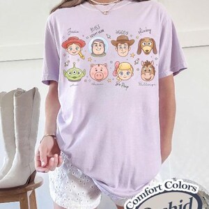 May include: A light purple t-shirt featuring cartoon character faces from the movie Toy Story. The shirt has the names of the characters written above their faces. The shirt also has the words "Comfort Colors" and "Orchid" printed on it.