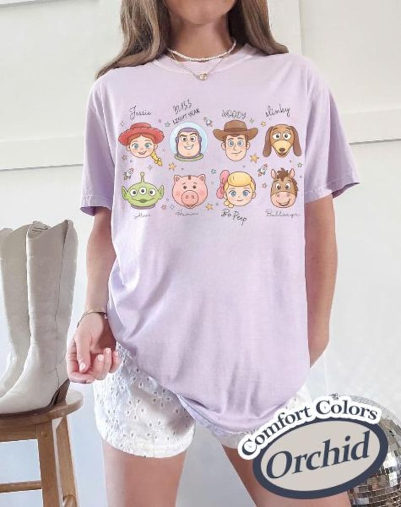 May include: A light purple t-shirt featuring cartoon character faces from the movie Toy Story. The shirt has the names of the characters written above their faces. The shirt also has the words "Comfort Colors" and "Orchid" printed on it.