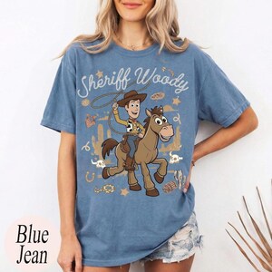 May include: A blue short-sleeve t-shirt featuring a graphic print of a cowboy character riding a brown horse. The text "Sheriff Woody" is printed above the character in a white script font, surrounded by small western-themed icons like stars.
