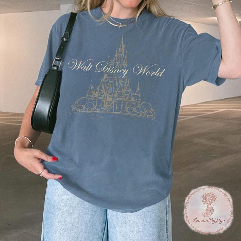 May include: Blue t-shirt with a gold illustration of a castle and the words "Walt Disney World". The shirt is a solid blue color and has short sleeves. The shirt is made of a soft material.