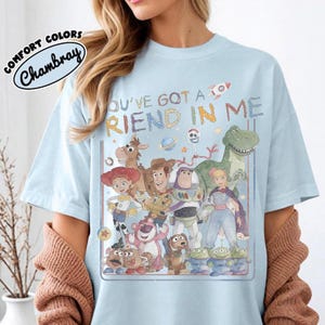 Pu&ograve; includere: Una t-shirt azzurra con una stampa grafica di vari personaggi di Toy Story, tra cui Woody, Buzz Lightyear, Jessie e Rex. Il testo sopra i personaggi recita YOU'VE GOT A FRIEND IN ME. Un logo nell'angolo riporta COMFORT COLORS Chambray.