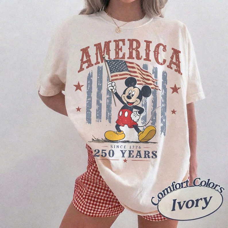 Pu&ograve; includere: T-shirt color avorio con grafica di Mickey Mouse con bandiera americana. La parola "AMERICA" &egrave; stampata sopra la grafica, con "250 YEARS" sotto. La maglietta &egrave; del marchio Comfort Colors.