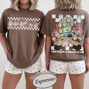 May include: Espresso-colored t-shirt featuring characters from Toy Story. The front has a checkered border with characters. The back displays more characters with the text "YOU'VE GOT A FRIEND IN ME". Paired with white shorts with a small floral pattern.