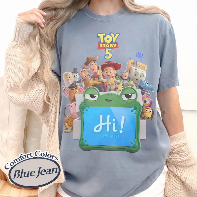 May include: A blue jean colored t-shirt featuring a Toy Story 5 graphic with various characters and a central green frog-shaped tablet displaying the text Hi! Let's play! The shirt includes a Comfort Colors Blue Jean label in the corner.