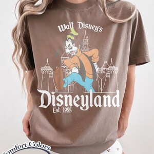 Puede incluir: Camiseta color espresso con un gr&aacute;fico vintage de Disneyland. El dise&ntilde;o incluye el texto "Walt Disney's Disneyland Est. 1955" con un personaje de dibujos animados e ilustraci&oacute;n de castillo. La camiseta es de Comfort Colors.