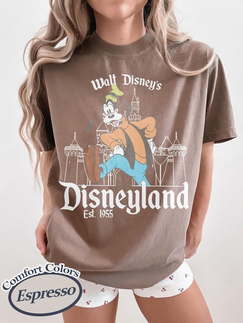 Puede incluir: Camiseta color espresso con un gr&aacute;fico vintage de Disneyland. El dise&ntilde;o incluye el texto "Walt Disney's Disneyland Est. 1955" con un personaje de dibujos animados e ilustraci&oacute;n de castillo. La camiseta es de Comfort Colors.