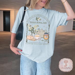 May include: Light blue t-shirt with a graphic featuring the text "WORLD FAMOUS GUMBO" and "TIANA'S PALACE". The design includes a teapot, flowers, honey, and the words "FLOWER TEA CAKE HONEY".
