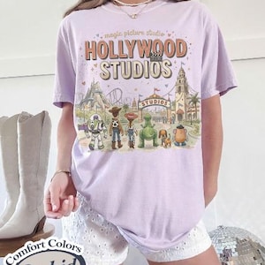 May include: A light purple t-shirt with a graphic of Hollywood Studios and various cartoon characters. The text "Hollywood Studios" is displayed in a vintage style. The shirt is a Comfort Colors Orchid.