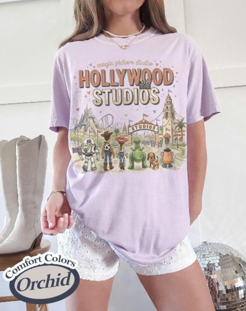 May include: A light purple t-shirt with a graphic of Hollywood Studios and various cartoon characters. The text "Hollywood Studios" is displayed in a vintage style. The shirt is a Comfort Colors Orchid.