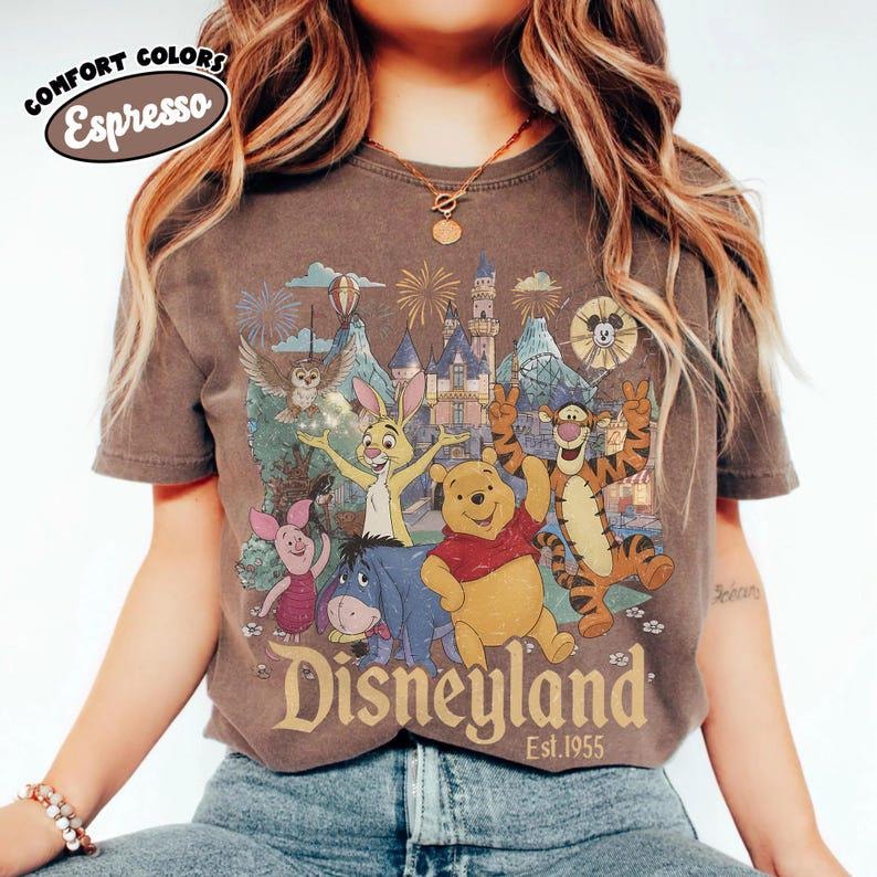 May include: An espresso-coloured short-sleeve t-shirt featuring a graphic print of Winnie the Pooh, Tigger, Piglet, Eeyore, and Rabbit in front of a castle. The text reads "Disneyland Est. 1955" and "COMFORT COLORS Espresso" is printed at the top.