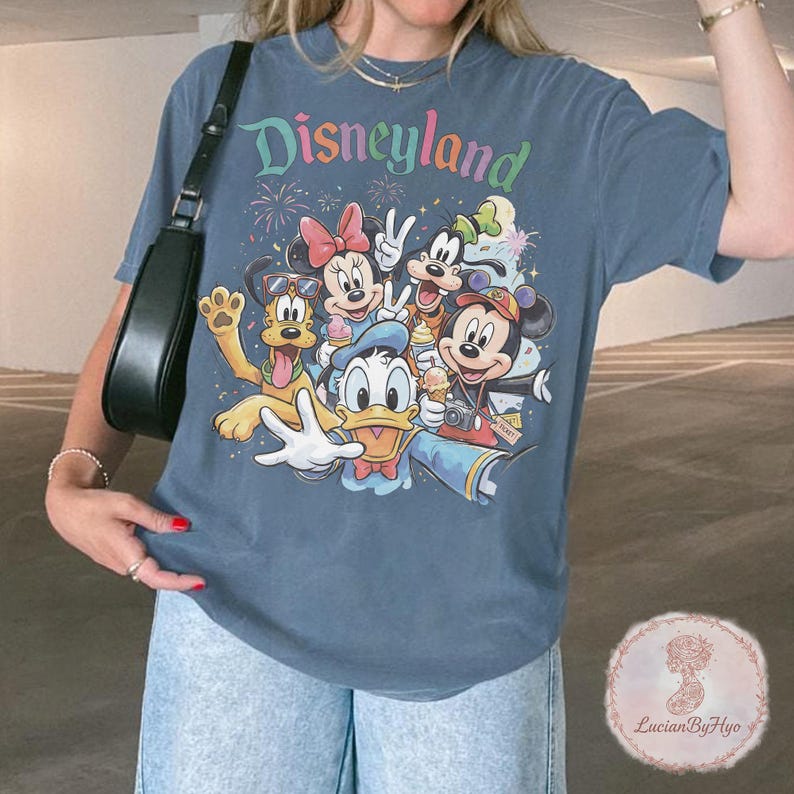 May include: Blue t-shirt featuring the word "Disneyland" in colorful text above a graphic of Disney characters including Mickey Mouse, Minnie Mouse, Donald Duck, and Pluto. The characters are smiling and waving.