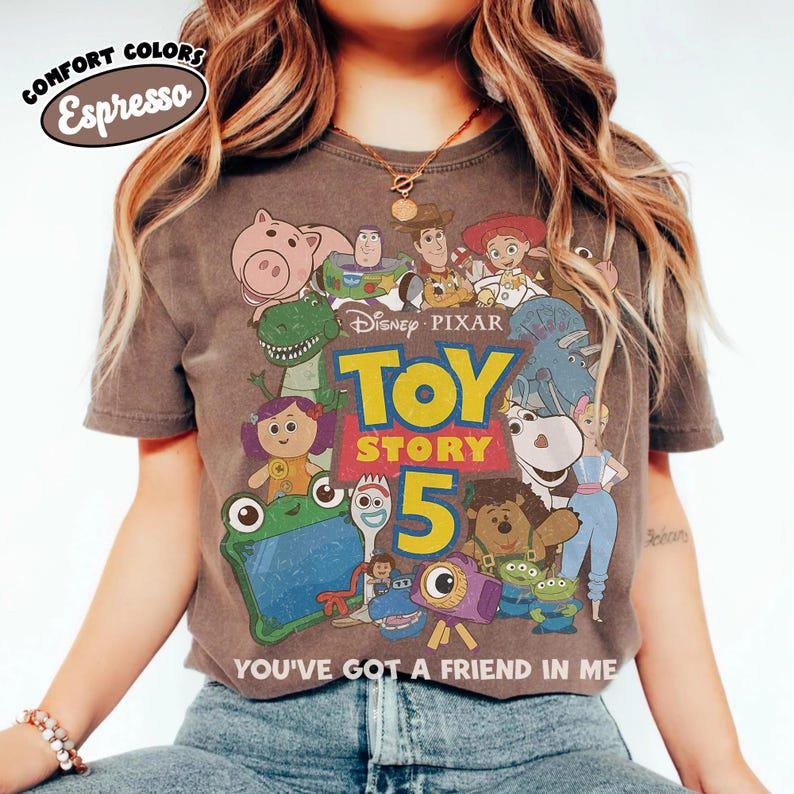 May include: An espresso-colored short-sleeve t-shirt featuring a graphic print of various Toy Story characters. The design includes the text Disney Pixar Toy Story 5 and the phrase You've Got A Friend In Me. The shirt has a crew neckline and a relaxed fit.