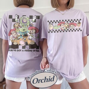 May include: Two lavender Comfort Colors t-shirts featuring Toy Story characters. One shirt has a large graphic on the back with the text "YOU'VE GOT A FRIEND IN ME." The other shirt has a smaller graphic on the front. Both shirts have a checkered pattern.