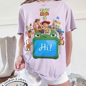 May include: A light purple t-shirt featuring a Toy Story 5 graphic with various characters above a green frog-shaped frame containing a blue screen with the text Hi! Let's play! The shirt is displayed with white eyelet shorts and light boots.