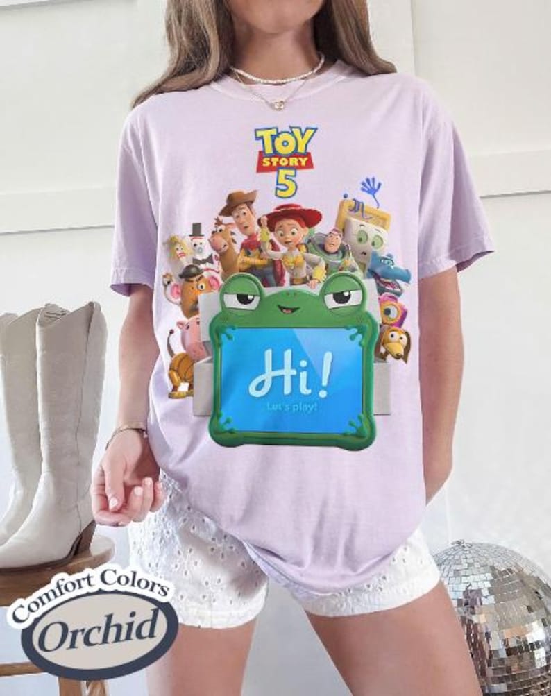 May include: A light purple t-shirt featuring a Toy Story 5 graphic with various characters above a green frog-shaped frame containing a blue screen with the text Hi! Let's play! The shirt is displayed with white eyelet shorts and light boots.