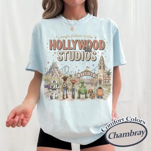 May include: Light blue t-shirt with a graphic of Hollywood Studios and various cartoon characters. The text "Hollywood Studios" is in a retro font. The shirt also features the text "Comfort Colors Chambray".