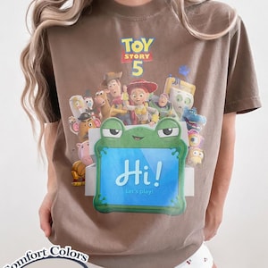 May include: A brown t-shirt featuring a graphic print of Toy Story 5 characters and a green frog-shaped tablet with the text Hi! Let's play! The shirt includes a Comfort Colors Espresso label in the corner, showcasing a crew neck and short sleeves.
