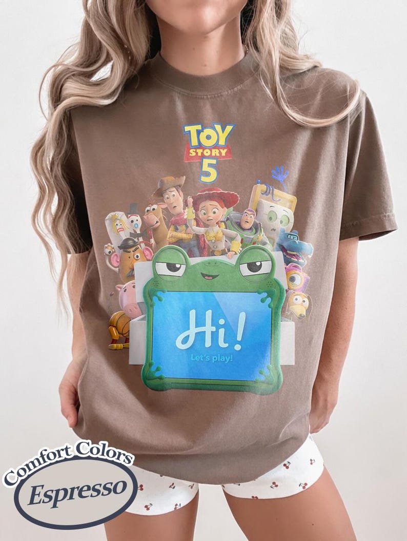 May include: A brown t-shirt featuring a graphic print of Toy Story 5 characters and a green frog-shaped tablet with the text Hi! Let's play! The shirt includes a Comfort Colors Espresso label in the corner, showcasing a crew neck and short sleeves.