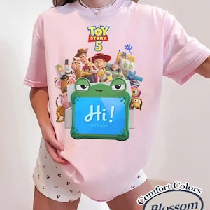 May include: A light pink t-shirt featuring a Toy Story 5 graphic with various characters and a green frog-shaped tablet displaying the text Hi! Let's play! The shirt is paired with white shorts featuring a small cherry print pattern throughout.