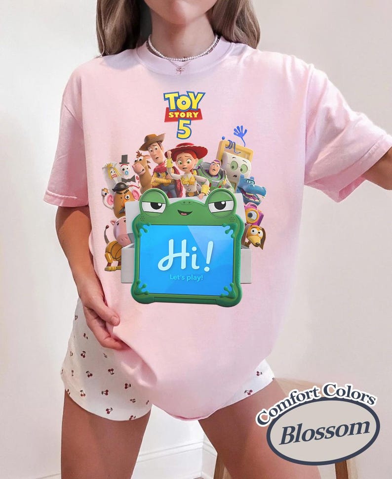 May include: A light pink t-shirt featuring a Toy Story 5 graphic with various characters and a green frog-shaped tablet displaying the text Hi! Let's play! The shirt is paired with white shorts featuring a small cherry print pattern throughout.