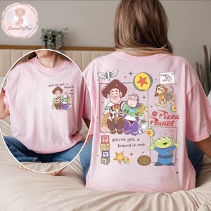 Puede incluir: Camiseta rosa claro con una ilustraci&oacute;n de dibujos animados de Toy Story. La parte delantera muestra a Woody y Buzz Lightyear con el texto "you've got a friend in me". La parte trasera presenta m&aacute;s personajes y el logo de Pizza Planet.