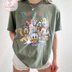 May include: Olive green t-shirt featuring the word "Disneyland" in colorful text above a graphic of Disney characters including Mickey Mouse, Minnie Mouse, Donald Duck, and Goofy. The shirt has a crew neck and short sleeves.