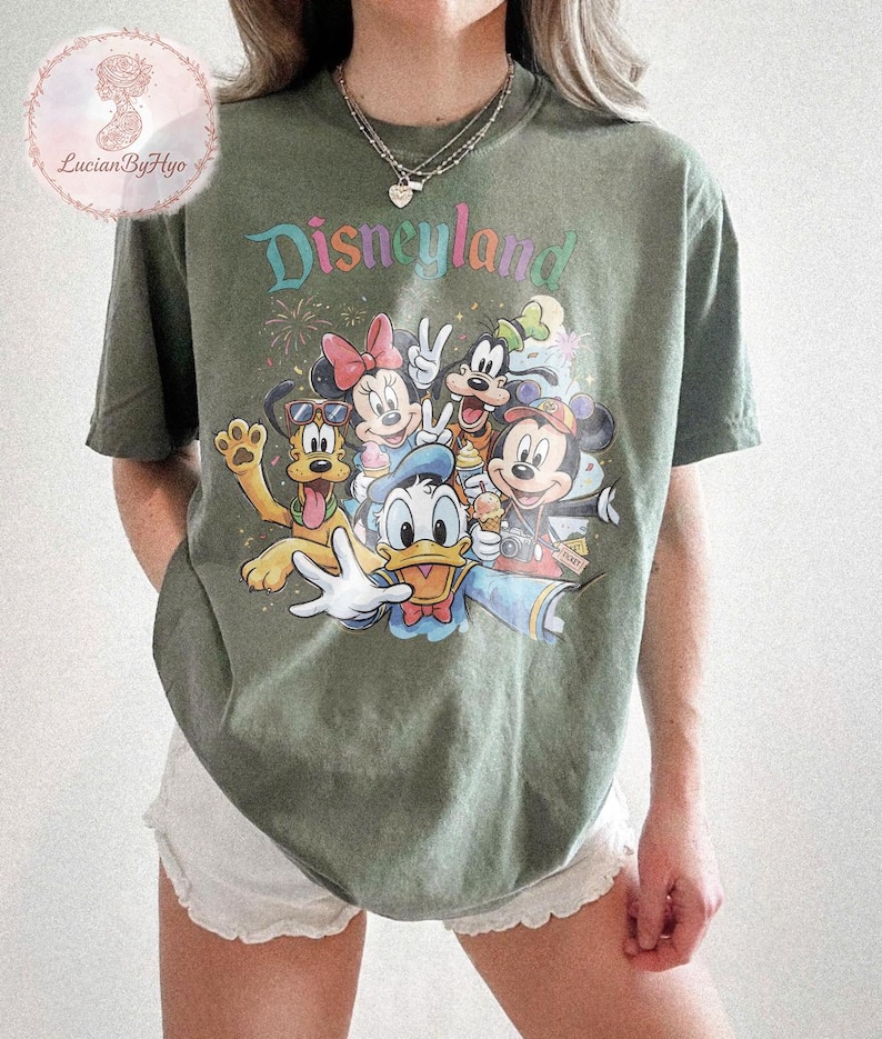 May include: Olive green t-shirt featuring the word "Disneyland" in colorful text above a graphic of Disney characters including Mickey Mouse, Minnie Mouse, Donald Duck, and Goofy. The shirt has a crew neck and short sleeves.