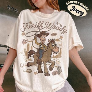 May include: An ivory t-shirt featuring a graphic of Sheriff Woody riding a horse. The design includes the text "Sheriff Woody" in a rope-style font, surrounded by desert elements like cacti, skulls, and stars. A "Comfort Colors Ivory" label is visible.