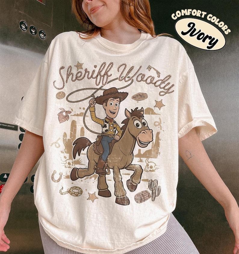 May include: An ivory t-shirt featuring a graphic of Sheriff Woody riding a horse. The design includes the text "Sheriff Woody" in a rope-style font, surrounded by desert elements like cacti, skulls, and stars. A "Comfort Colors Ivory" label is visible.