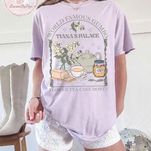 May include: A light purple t-shirt with a graphic featuring a teapot, teacup, honey jar, and flowers. Text reads "World Famous Gumbo Tiana's Palace" and "Flower Tea Cake Honey". The shirt is a comfortable, casual style.