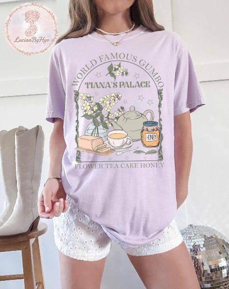 May include: A light purple t-shirt with a graphic featuring a teapot, teacup, honey jar, and flowers. Text reads "World Famous Gumbo Tiana's Palace" and "Flower Tea Cake Honey". The shirt is a comfortable, casual style.