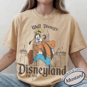 Puede incluir: Camiseta color mostaza con Goofy frente al castillo de Disneyland. La camiseta tiene el texto "Walt Disney's Disneyland Est. 1955" en un estilo vintage. La camiseta es parte de la colecci&oacute;n Comfort Colors.