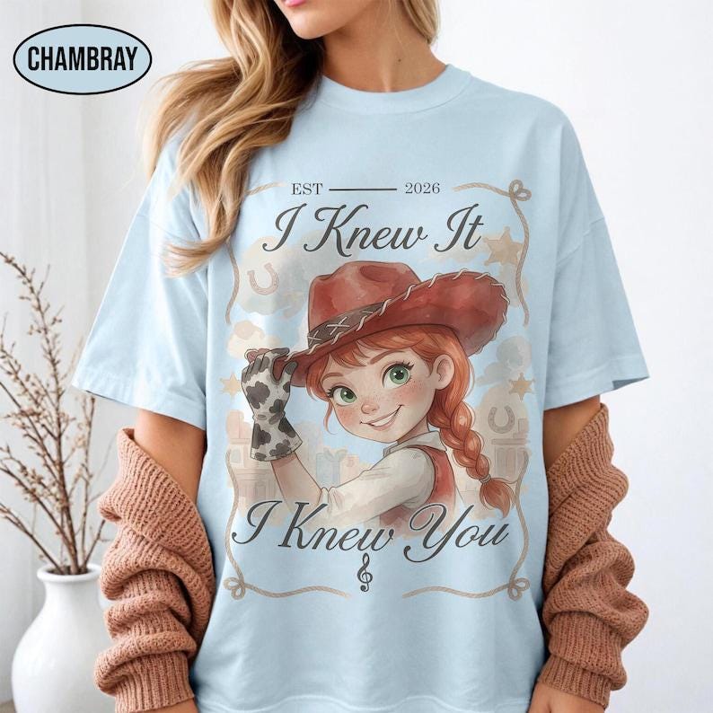 May include: A light blue t-shirt featuring a graphic of a cartoon cowgirl with red hair in braids and a brown cowboy hat. The text reads EST 2026, I Knew It, and I Knew You. The design includes a lasso border, stars, and horseshoes.