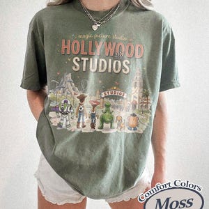 May include: Moss green t-shirt with a graphic of Hollywood Studios and Disney characters. The shirt features the text "Hollywood Studios" and "magic picture studio" in a vintage style. The shirt is made of Comfort Colors material.
