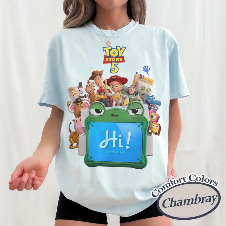 May include: A light blue t-shirt featuring a Toy Story 5 graphic with various characters and a green frog-shaped tablet displaying the text Hi! Let's play! The shirt has a crew neckline and short sleeves, showcasing a colorful print on the front.