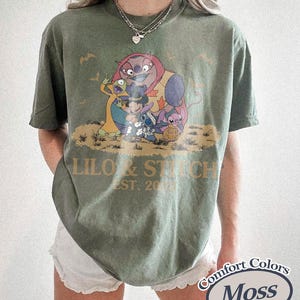 K&ouml;nnte beinhalten: Moosgr&uuml;nes T-Shirt mit einer Grafik von Lilo & Stitch-Charakteren und dem Text "Lilo & Stitch EST. 2002". Das Shirt ist von der Marke Comfort Colors.