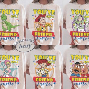 Op de afbeelding: Ivoor kleurige T-shirts met verschillende Toy Story-personages, waaronder Rex, Woody, Buzz Lightyear, Bo Peep, Jessie en Mr. en Mrs. Potato Head. Elk shirt heeft de tekst "YOU'VE GOT A FRIEND IN ME!" in geel en rood.