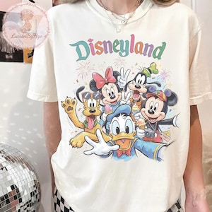 May include: Cream-colored t-shirt featuring the word "Disneyland" in colorful letters above a graphic of Disney characters, including Mickey Mouse, Minnie Mouse, Donald Duck, Goofy, and Pluto. The characters are smiling and holding ice cream cones.