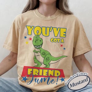 Op de afbeelding: Mosterdkleurig T-shirt met een groene cartoon-dinosaurus en de tekst "YOU'VE GOT A FRIEND" in geel en blauw. Het woord "June!" staat in blauwe letters onder de dinosaurus.