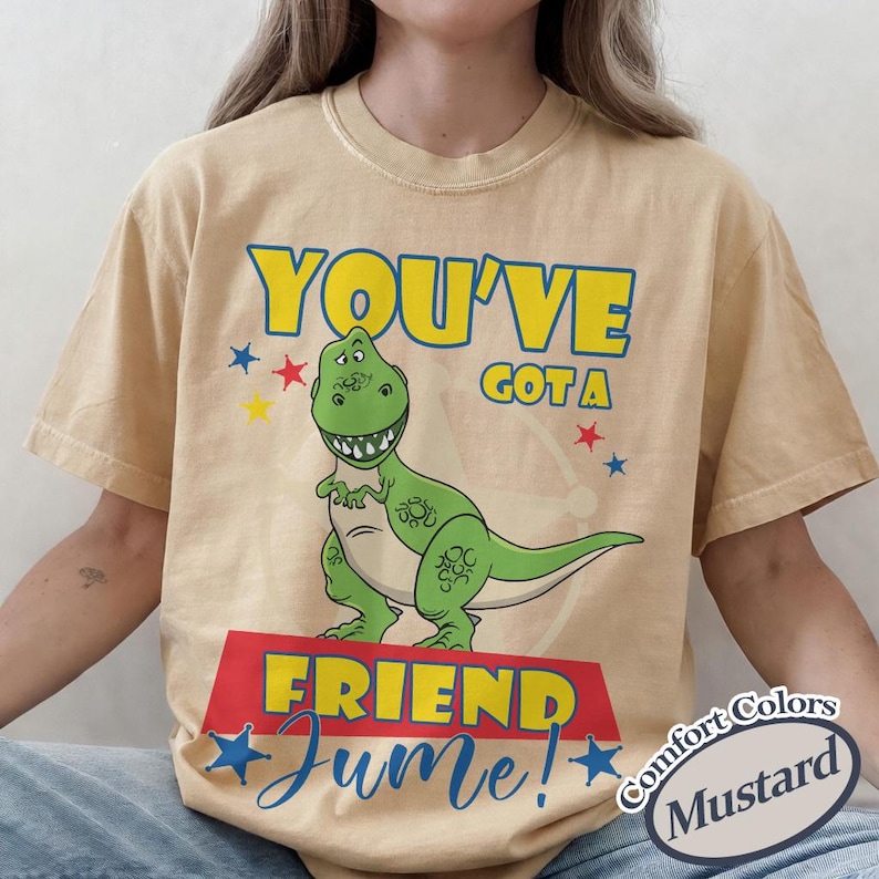 Op de afbeelding: Mosterdkleurig T-shirt met een groene cartoon-dinosaurus en de tekst "YOU'VE GOT A FRIEND" in geel en blauw. Het woord "June!" staat in blauwe letters onder de dinosaurus.