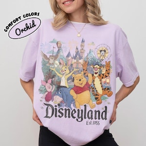 May include: A light purple t-shirt featuring a graphic print of Winnie the Pooh, Piglet, Tigger, Eeyore, and Rabbit in front of a castle. The text reads "Disneyland Est. 1955" and "COMFORT COLORS Orchid". The design includes fireworks and a Ferris wheel.
