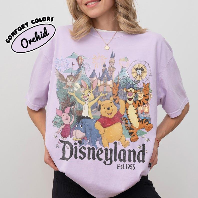 May include: A light purple t-shirt featuring a graphic print of Winnie the Pooh, Piglet, Tigger, Eeyore, and Rabbit in front of a castle. The text reads "Disneyland Est. 1955" and "COMFORT COLORS Orchid". The design includes fireworks and a Ferris wheel.