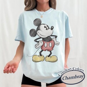 May include: Light blue t-shirt featuring a sketch-style Mickey Mouse graphic. The cartoon character is depicted in black, red, and yellow. The shirt has short sleeves and a crew neck. The text "Comfort Colors Chambray" is printed on the bottom right.