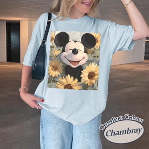 Puede incluir: Camiseta azul claro con un dise&ntilde;o de Mickey Mouse sonriente rodeado de girasoles. El gr&aacute;fico est&aacute; centrado en la camiseta. La camiseta tiene mangas cortas y un corte holgado. El texto "Comfort Colors Chambray" est&aacute; impreso en la parte inferior derecha.
