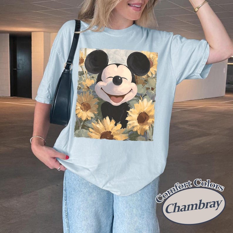 Puede incluir: Camiseta azul claro con un dise&ntilde;o de Mickey Mouse sonriente rodeado de girasoles. El gr&aacute;fico est&aacute; centrado en la camiseta. La camiseta tiene mangas cortas y un corte holgado. El texto "Comfort Colors Chambray" est&aacute; impreso en la parte inferior derecha.