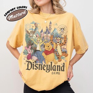May include: A mustard yellow t-shirt featuring a graphic print of Winnie the Pooh, Tigger, Piglet, Eeyore, and Rabbit in front of a castle. The text reads "Disneyland Est. 1955" in a dark font. A "Comfort Colors Mustard" label is in the top left corner.